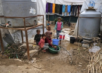 Children sit by as a woman washes dishes in a plastic basin outside a tent at a make-shift camp for Syrian refugees in Talhayat in the Akkar district in north Lebanon on October 26, 2022. (Photo by Ibrahim Chalhoub / AFP)